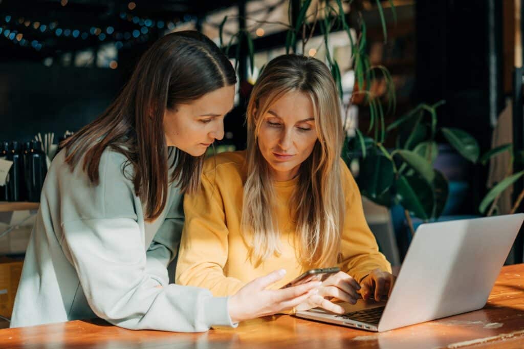 Two business women discuss online content and use phone and laptop.