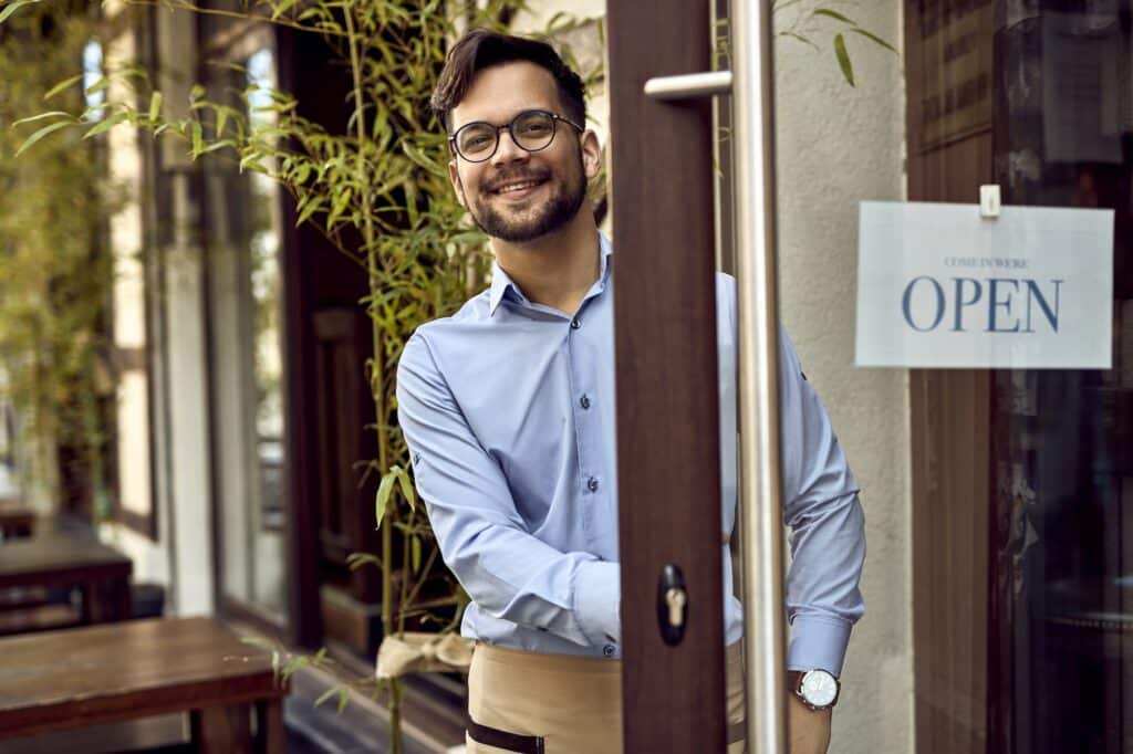 Young happy waiter standing at cafe's front door.