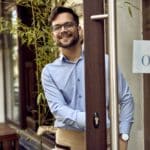 Young happy waiter standing at cafe's front door.